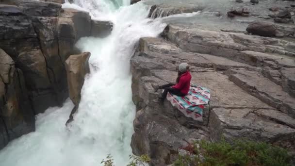 女性は寒い秋の日にカナダの田舎の滝と氷河川の上の岩の上に座っています Sunwapta Falls ジャスパー国立公園 カナダ — ストック動画