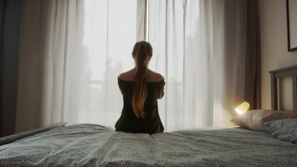 Beautiful woman with long hair gets out of bed and opens the bedroom curtains in the morning
