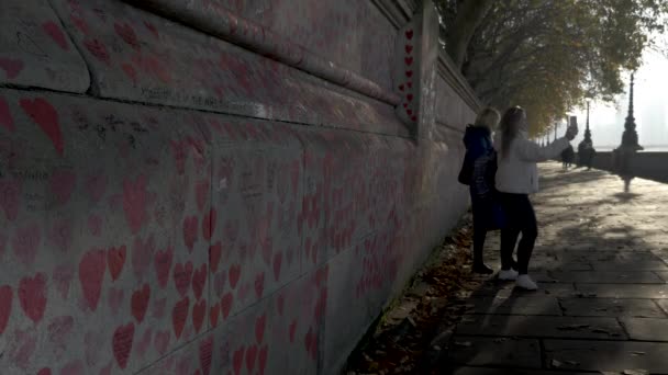 彼女の携帯電話上のライブビデオ通話で友人に話して国立コヴィド記念館の壁の前に立っている2人の若い女性 壁に手で描かれたピンクの心でいっぱい ロンドン イギリス — ストック動画