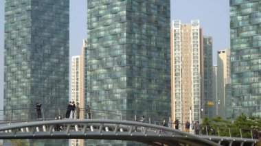 Incheon, Güney Kore 'deki Songdo Central Park' a giden yaya köprüsü, yüz maskeleri takan insanlar ve arka planda modern cam gökdelen, Incheon Güney Kore