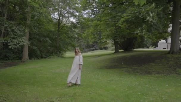 怀孕的年轻女子穿着漂亮的白色连衣裙穿过绿油油的花园 这是生育照的概念 — 图库视频影像