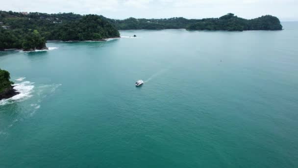 濃い緑の雨林に覆われた岩の海岸線の近くのターコイズブルーの海で航行する船の空中軌道 Naohomi Park Costa Rica — ストック動画