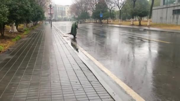 中国の伝統的な中国のほうきで雨の日に 緑のレインコートを着て大人の高齢者の歩道上の歩行ショットは 空の濡れた孤独な通りを掃引します — ストック動画