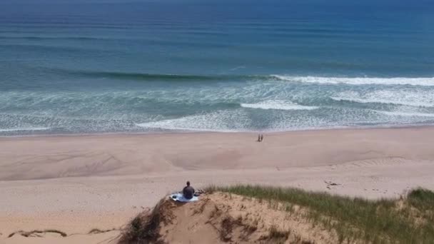 巨大な砂の砂丘の上に座っている観光客を飛び越えるドローンは大西洋の波を明らかに — ストック動画