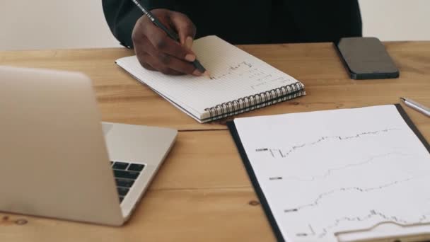 Detailní záběr obchodníka, jak si dělá poznámky do papíru s akciemi, který sedí před laptopem a grafy. Bez tváře.