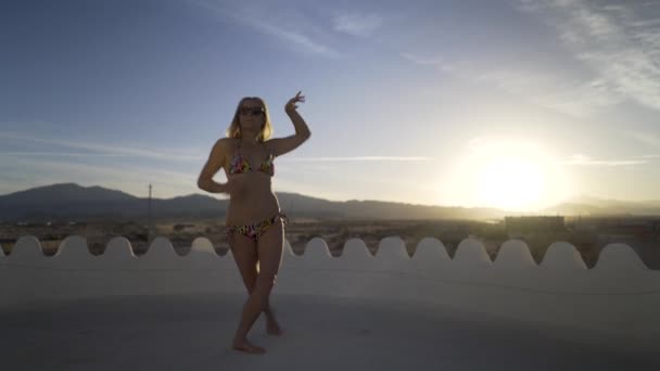 Exotic belly dancer with sunglasses and bikini set against a blue sky and mountains and setting sun does a body wave.
