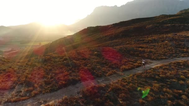日没時に南アフリカの山を下る未舗装の道を走る男の広い空中ショット — ストック動画