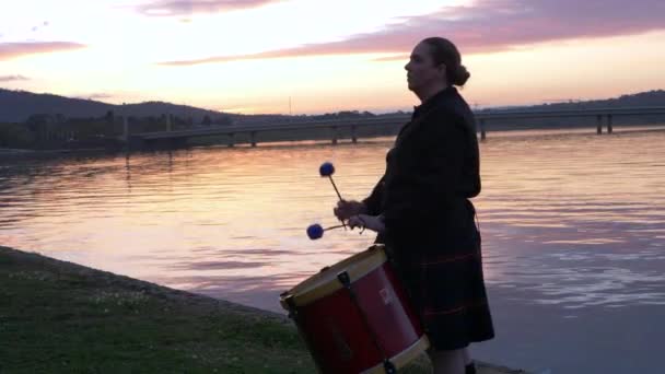 マーチングバンドのメンバーである女性が 湖のそばの秋の日の早朝の日の出にテナントドラムを演奏しています 背景のシーンは美しい日の出の空と湖です — ストック動画