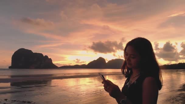 かわいいアジアの女の子は カラフルな夕日の背景と島のシルエットに笑みを浮かべてビーチで携帯電話を使用しています アジア Koh Phangan Island Thailand — ストック動画