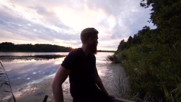 一个留着大胡子的大男子主义男子在美丽的落日下坐在湖边 让人叹为观止的水 定格滑翔机镜头以慢动作从模型上移开 — 图库视频影像