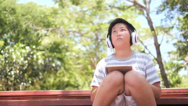 屋外の公園で座ってヘッドホンで陽気なアジアのプレティーンの少年 — ストック動画