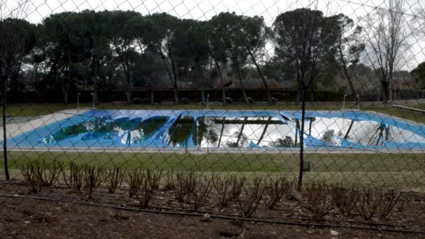 Široký záběr velkého krytého bazénu přes drátěný plot. Na vršku plachty jsou vidět odrazy na vodě.