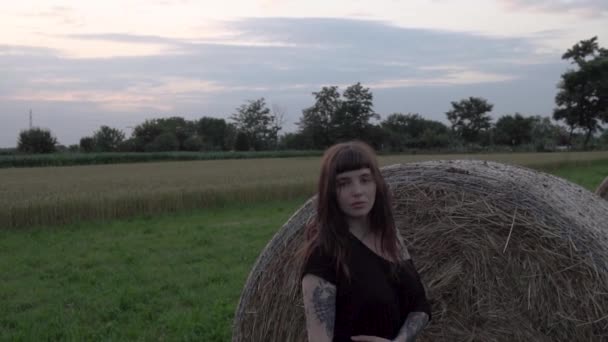 美しい少女はヨーロッパの日没時に農場の干し草の大きなベールの近くに立つ — ストック動画