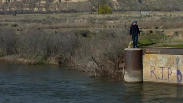 青いジャケットの男が川に何か投げ込んでいる 川は錆びた鉄橋のふもとで静かに流れている — ストック動画