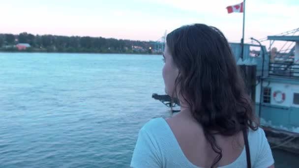 夕阳西下时 坐在木板路上看着弗雷泽河的年轻女子的近视 加拿大国旗在后方飘扬 加拿大新威斯敏斯特码头 — 图库视频影像