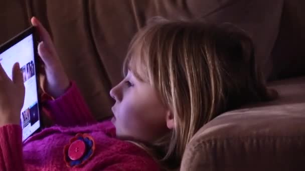 若いブロンドの女の子はタブレットに集中しソファに横になり — ストック動画