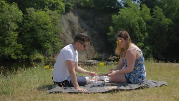 小河边僻静地区的一对年轻夫妇坐在毛毯上野餐 — 图库视频影像