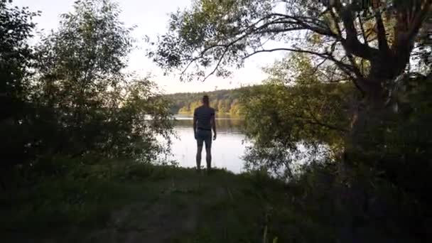 スウェーデンのストックホルムにある美しい湖のそばに背の高い白人男性が立っています 夏の夜 モデル緑の木々に囲まれて モデルに向かって動く安定したグライドカム映像 穏やかな屋外エリア — ストック動画