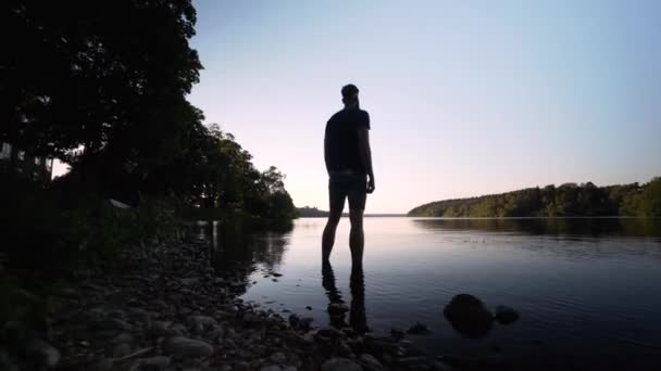 スウェーデンのストックホルムにある湖に背の高い白人男性が立っている 美しい夏の夜 青空と暖かい外 モデルから離れた安定したグライドカム映像 水の上の美しい反射 — ストック動画