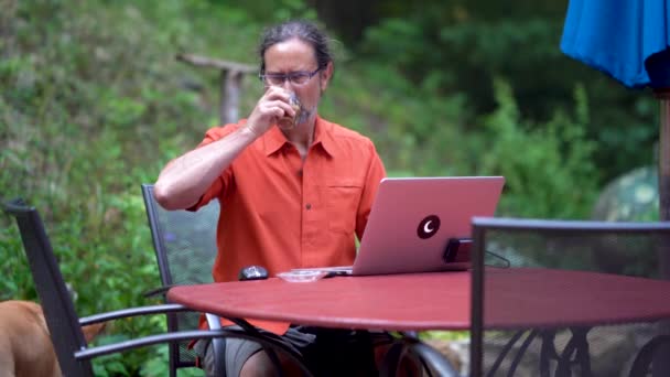 一个成熟的男人坐在花园的室外咖啡馆里喝咖啡 和狗一起在电脑上工作 — 图库视频影像