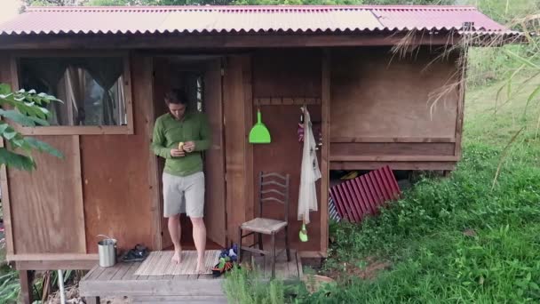 Muž Kráčí Ven Loupá Banán Ráno Před Plechovou Střešní Boudou — Stock video