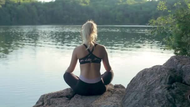 Fiatal nő jógázik a tónál naplementekor. Ülő jóga pózok.