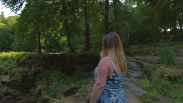 短い青いドレスの女性は緑豊かな緑の中で小さな川の岩のオーバーハングに向かって歩く — ストック動画