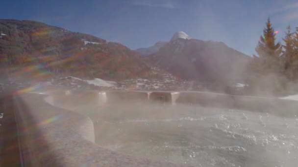 一个炎热的浴缸泡在阳光下 后面是白雪覆盖的山丘和一个滑雪胜地 摄像机从左向右滑动 — 图库视频影像