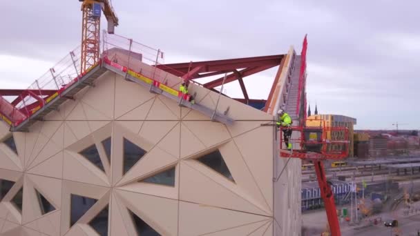 建設中の建物の上部付近に足場の組み立てを完了する建設労働者の空中映像 曇り空と周囲の街の景色 — ストック動画