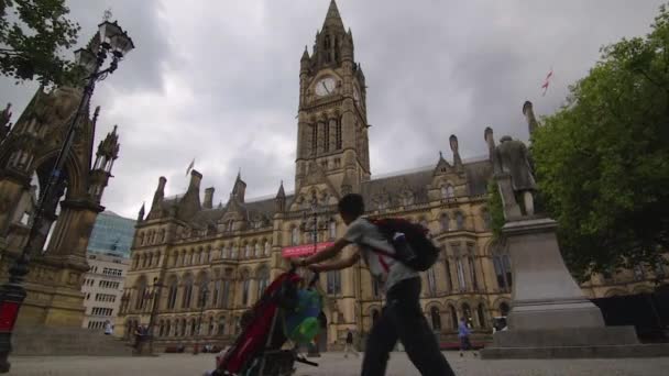 Pohled na budovu Manchesterské radnice s kolemjdoucími v popředí a mužem tlačícím dětský kočárek
