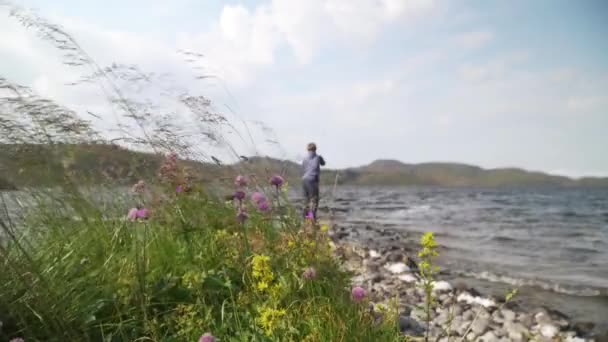風に揺れる美しい紫色と黄色の野生の花や草の閉鎖 マスとサーモンの釣竿を背景に女性が立っている — ストック動画