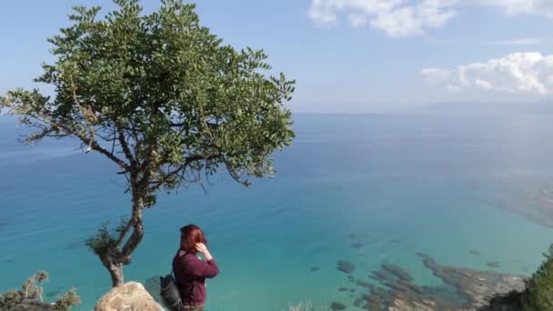 一个女人站在一座山边的一棵树旁 凝视着美丽的蓝色海水 — 图库视频影像