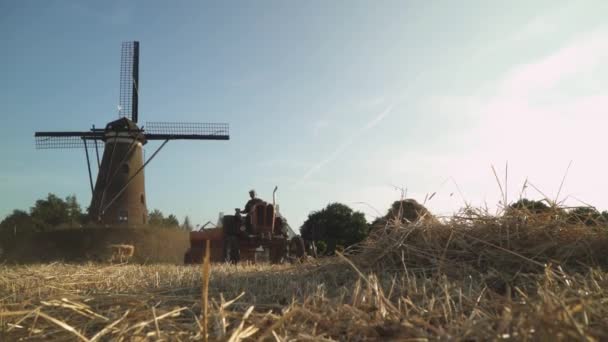 干し草の俵を生産する干し草のプレスとヴィンテージトラクター 背景に風車があります 典型的な ロマンチックな 絵のようなオランダのシーン 低角度 — ストック動画