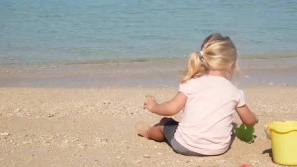 坐在沙滩上的金发小女孩在沙滩上玩水 — 图库视频影像