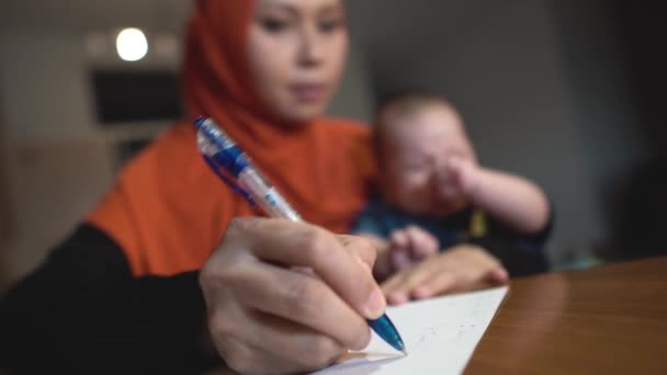 紙にペンで書いている間に机の上に座って泣いて赤ちゃんとFocusイスラム教徒の母親から — ストック動画