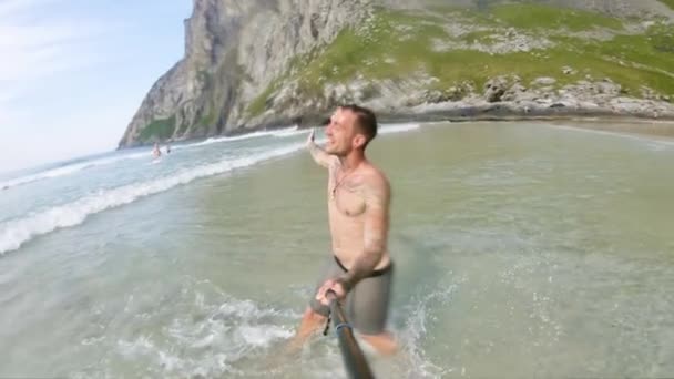 笑顔の男が波のようにカメラを持って海を歩いている 海と周囲のスローモーションビュー 深く岩の多い山が水の端から立ち上がり — ストック動画