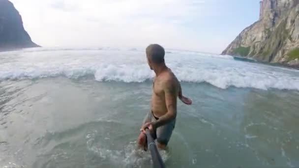 海を楽しむ男は冷たい波が彼の周りを洗うことに驚いている 彼は冷たい水の温度に反応する この砂の入り江の周りの背景には 海から険しい岩肌が隆起しています — ストック動画