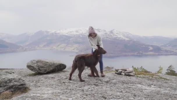 Woman with glasses, red cap, grey sweater, blue jeans and brown boots playing and petting a chocolate flat coated retriever on the top of a mountain with a view to the fjord and snowy mountains