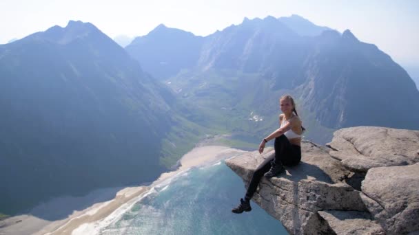 Slow motion view of a woman sitting on a mountain ledge overlooking the beautiful blue water of a fjord far below as she turns away from the camera. Mountain scenery, a deep green valley and beach.
