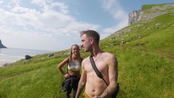 若いカップルは暖かい日差しを楽しむKvalvikaビーチに向かってRyten山を下ります カメラに向かって声を上げながら歩き回っている男 — ストック動画