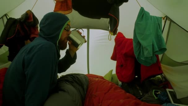 嵐の中テントの中で寝袋で男ハイカーが待って酒を飲んでテントの壁が風の突風で揺れ — ストック動画