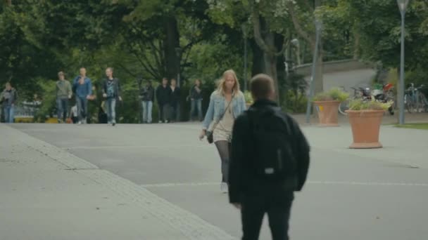 Student with backpack walks down the street away from the camera as a young blonde woman in a denim jacket approaches.