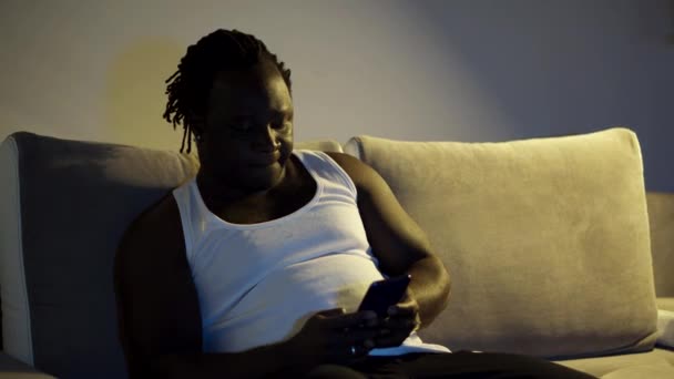 身穿背心的黑人坐在沙发上 用手机发短信 晚上换电视频道 — 图库视频影像