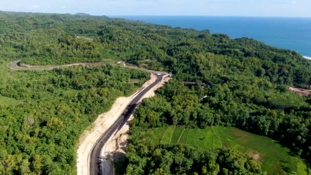インドネシアの活気ある森林地帯で建設中の青い海の水と曲がりくねった道 空中ビュー — ストック動画