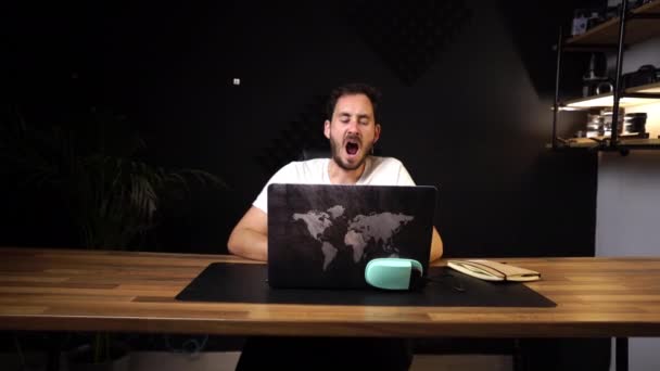 若い白人男性は 世界中の新しい旅行冒険を計画している世界の地図と彼のラップトップで働いている彼の現代的なスタジオに座っている間Yawning — ストック動画