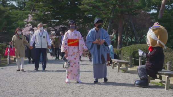 兼六園を歩く若いアジア系日本人の仮面カップルと地元のマスコットライタンに手を振る — ストック動画