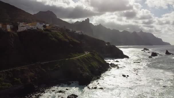 Aerial drone view of a rugged mountainous Atlantic ocean coastline with a paved road and a small village on top of it. Recorded in Parque Rural de Anaga in Northern Tenerife in the Canary Islands.
