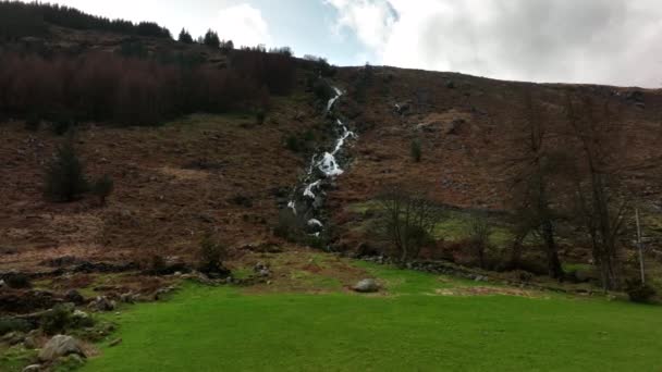 Carawaystick Waterfall, Glenmalure, Wicklow, Irsko, únor2022. Dron se tlačí přes zelené pole k úpatí vodopádů a postupně se naklání dolů.