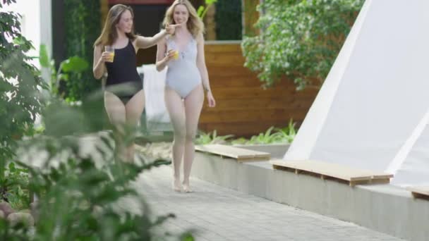 一对迷人的性感健康的女孩穿着一件泳衣 喝着一杯排毒果汁 一起走在一个现代豪华度假胜地的温泉里享受假期 — 图库视频影像