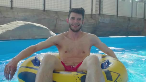 年轻快乐的高加索男子坐在充气救生圈划船在游泳池水慢动作的形象有趣的暑假 — 图库视频影像